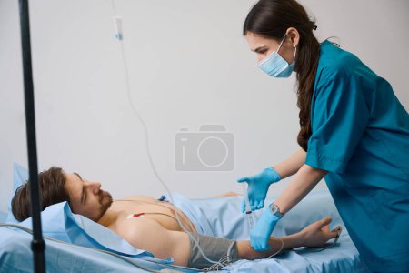 Téléchargez les photos : Une jeune infirmière injecte des médicaments dans un goutte à goutte, un homme est connecté à un holter - en image libre de droit