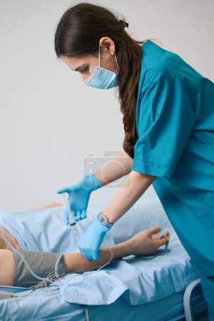 Foto de Enfermera en una máscara protectora inyecta medicina en un goteo, un hombre se acuesta en una cama de hospital - Imagen libre de derechos