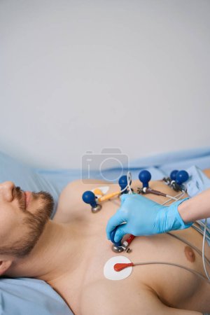Téléchargez les photos : Cardiologue dans des gants de protection attache les ventouses du cardiographe à un patient, un homme se trouve sur un lit d'hôpital - en image libre de droit