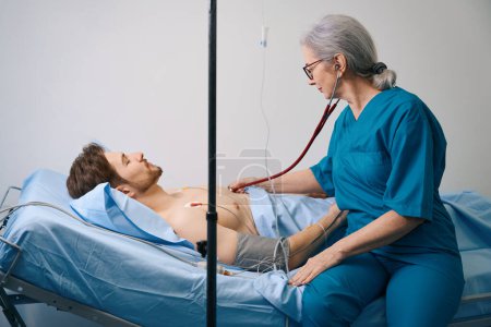 Foto de Un hombre barbudo yace bajo un goteo con un torso desnudo, un médico anciano lo escucha con un estetoscopio - Imagen libre de derechos