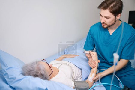 Freundliche Krankenschwester hält die Hand einer Patientin, die auf dem Bett liegt, er misst ihren Puls