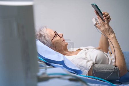 Foto de Mujer optimista está enviando mensajes de texto en un teléfono móvil desde una habitación de hospital, ella está conectada a un monitor de paciente - Imagen libre de derechos