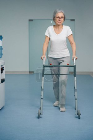 Téléchargez les photos : Belle femme âgée se déplace avec un marcheur le long du couloir de l'hôpital, une femme en vêtements confortables - en image libre de droit