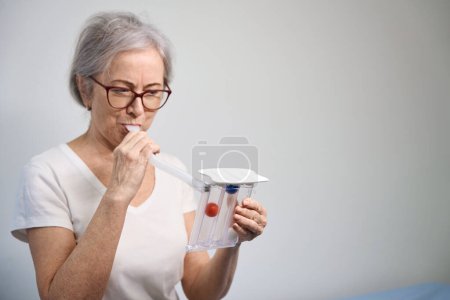 Téléchargez les photos : Patiente âgée gonfle ses poumons après la chirurgie sur un simulateur spécial, une femme en vêtements confortables - en image libre de droit