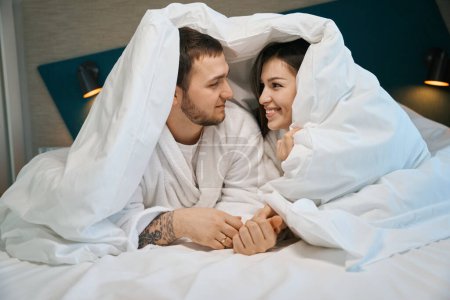Téléchargez les photos : Jeune mari et femme bavardent joyeusement sous les couvertures sur le grand lit, les gars en peignoirs doux - en image libre de droit