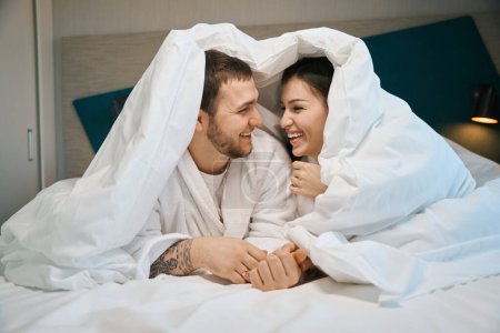 Téléchargez les photos : Les jeunes conjoints s'amusent à bavarder sous les couvertures sur un grand lit, les gars en peignoirs doux - en image libre de droit