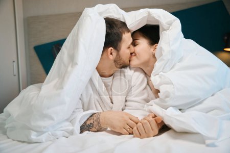 Téléchargez les photos : Jeune mari et femme embrasser mignon sous les couvertures sur le grand lit, les gars en peignoirs doux - en image libre de droit