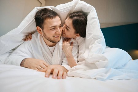 Téléchargez les photos : Maris amoureux étreignant et chuchotant sous les couvertures sur un grand lit, les gars en peignoirs doux - en image libre de droit