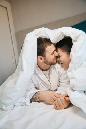 Téléchargez les photos : Charmant mari et femme rient heureux sous les couvertures sur un grand lit, les gars en peignoirs doux - en image libre de droit