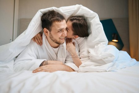 Téléchargez les photos : Joyeux conjoints câlins et chuchoter sous les couvertures sur un grand lit, les gars en peignoirs doux - en image libre de droit