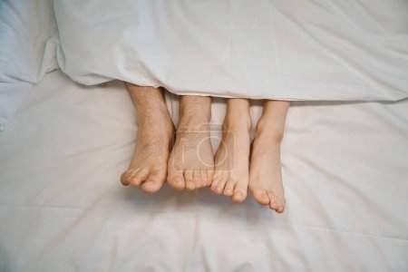 Foto de Dos pares de piernas jóvenes, un macho y una encantadora hembra, asoman desde debajo de las sábanas, la pareja disfruta de sus vacaciones - Imagen libre de derechos