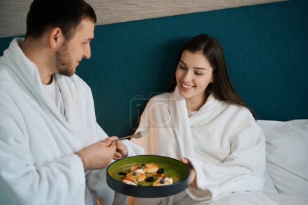 Foto de Joven en la cama da un tenedor a su esposa, la pareja se encuentra con el desayuno en el dormitorio - Imagen libre de derechos