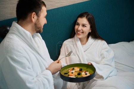 Téléchargez les photos : Jeune homme traite sa femme au petit déjeuner dans la chambre, le couple est situé sur un grand lit - en image libre de droit