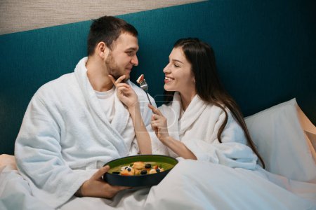 Téléchargez les photos : Femme attentionnée traite son mari au petit déjeuner dans la chambre, le couple est situé dans un grand lit - en image libre de droit