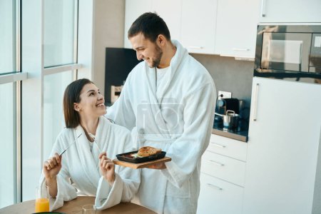 Téléchargez les photos : Amour jeune mari traite sa charmante femme pour le petit déjeuner dans une atmosphère chaleureuse, les gars en peignoirs confortables - en image libre de droit