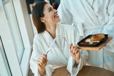 Téléchargez les photos : Homme traite sa charmante femme avec un petit déjeuner traditionnel dans une atmosphère chaleureuse, couple est situé à grande fenêtre de vue - en image libre de droit