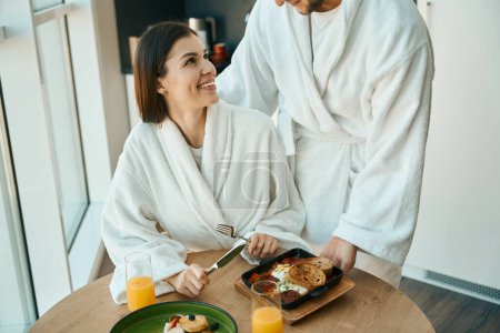 Téléchargez les photos : Jeune mari attentionné traite sa douce femme au petit déjeuner dans une atmosphère chaleureuse, les gars en peignoirs confortables - en image libre de droit