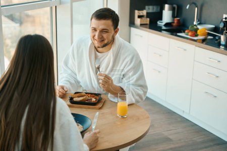 Téléchargez les photos : Agréable jeune couple bavarder au petit déjeuner dans une atmosphère chaleureuse, les gars en peignoirs doux - en image libre de droit