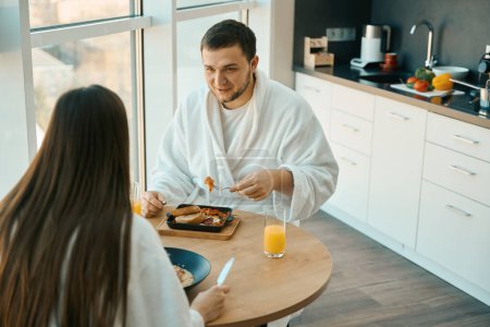 Téléchargez les photos : Les jeunes conjoints bavardent bien pendant le petit déjeuner dans une atmosphère chaleureuse, les gars en peignoirs doux - en image libre de droit