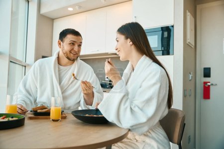 Téléchargez les photos : Joyeux jeune couple prenant le petit déjeuner dans un environnement confortable, les gars en peignoirs doux - en image libre de droit