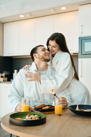 Téléchargez les photos : Joyeux jeune couple en peignoirs prenant le petit déjeuner dans une cuisine moderne confortable, une femme étreint doucement un homme - en image libre de droit