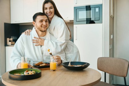 Foto de Alegre pareja joven en albornoces desayunando en una acogedora cocina minimalista, una mujer abraza suavemente a un hombre - Imagen libre de derechos
