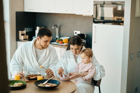 Téléchargez les photos : Mignonne idylle familiale par un matin ensoleillé, une jeune famille prend le petit déjeuner à la table de la cuisine - en image libre de droit