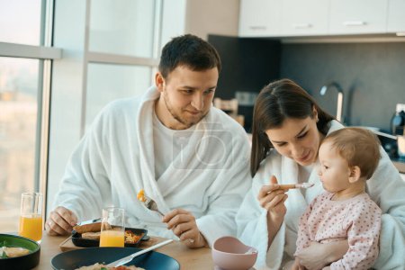 Photo for Joyful beautiful woman is feeding her little daughter, mom, dad and a small child are having breakfast - Royalty Free Image
