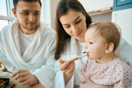 Photo for Happy cute mom feeds her little daughter, at breakfast mom, dad and little baby - Royalty Free Image