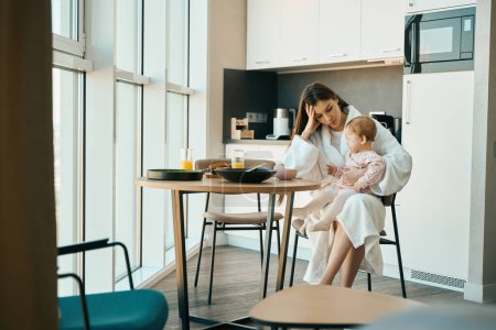 Photo for Upset woman in bathrobe sits at the kitchen table near the view window, she has crying baby in her arms - Royalty Free Image