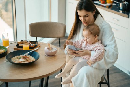 Téléchargez les photos : Charmante mère s'assoit avec sa petite fille à la table de la cuisine, la femme montre quelque chose sur le bébé au téléphone - en image libre de droit