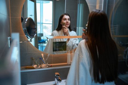 Foto de Morena joven de pelo largo se para frente al espejo en el baño y se da un masaje facial con un rodillo de jade - Imagen libre de derechos