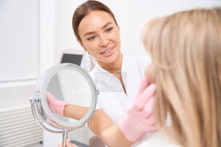 Foto de Cliente de la clínica de medicina estética en una consulta con un cosmetólogo, una mujer se mira en el espejo - Imagen libre de derechos
