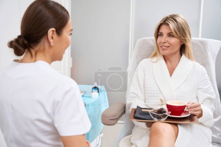 Foto de Cosmetólogo médico lleva a cabo una cita en una clínica de medicina estética, el paciente está sentado cómodamente con té y fruta - Imagen libre de derechos