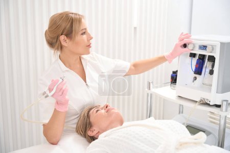 Téléchargez les photos : Jeune cosmétologue ajuste le laser CO2 pour une opération de refaçage de la peau, le patient s'allonge sur un canapé de cosmétologie - en image libre de droit