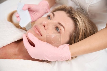 Téléchargez les photos : Patient subit une procédure innovante de nettoyage du visage par ultrasons, le spécialiste utilise un nettoyeur à ultrasons - en image libre de droit