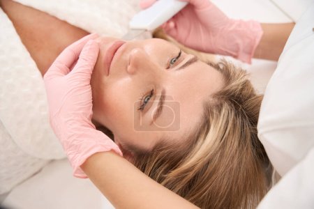 Foto de Mujer en procedimiento de limpieza facial ultrasónica de bienestar, especialista utiliza fregador ultrasónico - Imagen libre de derechos
