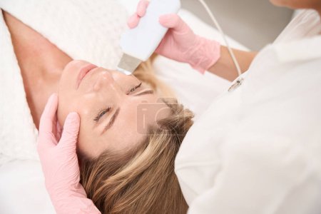Foto de Mujer en procedimiento de limpieza facial ultrasónica antienvejecimiento, especialista utiliza fregador ultrasónico - Imagen libre de derechos