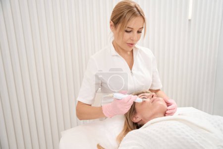 Foto de Especialista cosmetólogo realiza el procedimiento de limpieza ultrasónica de la cara, el paciente en un sofá de cosmetología - Imagen libre de derechos