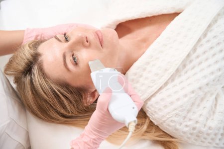 Téléchargez les photos : Femme sur la procédure de nettoyage du visage par ultrasons, la patiente repose sur un canapé de cosmétologie - en image libre de droit