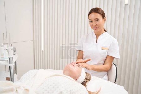 Foto de Mujer esteticista hace un masaje facial a un paciente en un albornoz, un empleado en un uniforme médico - Imagen libre de derechos