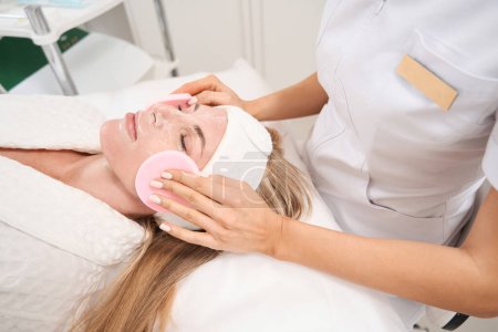 Foto de Cliente en una sesión de procedimientos de atención en la clínica de medicina estética, un cosmetólogo retira la máscara con esponjas especiales - Imagen libre de derechos