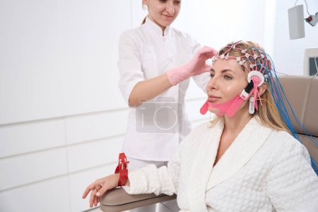 Photo for Young employee of a medical center attaches a cap with electrodes to a womans head for a diagnostic procedure - Royalty Free Image
