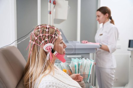 Téléchargez les photos : Diagnosticien dans un centre de réadaptation effectue une procédure d'électroencéphalogramme à une jeune patiente dans un bouchon avec des électrodes - en image libre de droit