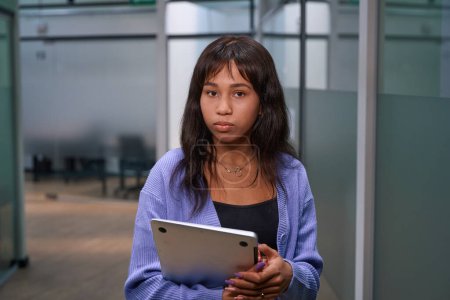 Téléchargez les photos : Jeune travailleuse regardant la caméra alors qu'elle se tenait debout avec un ordinateur portable dans ses mains dans un couloir étroit - en image libre de droit