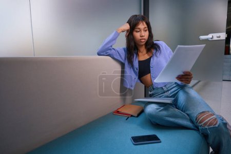 Téléchargez les photos : Jolie femme en vêtements élégants se détendre sur le canapé tout en regardant les papiers dans la chambre moderne - en image libre de droit