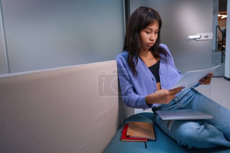 Téléchargez les photos : Vue latérale de la femme responsable assise sur un canapé confortable et travaillant avec des papiers dans un bureau moderne - en image libre de droit