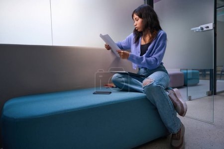 Téléchargez les photos : Jeune femme portant des vêtements fantaisie assis sur un canapé confortable et des notes d'examen dans la chambre moderne - en image libre de droit