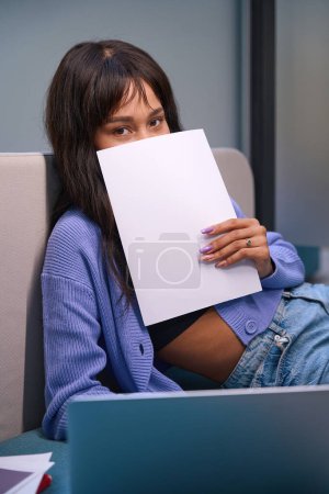 Téléchargez les photos : Jeune femme regardant la caméra et couvrant la moitié de son visage avec du papier tout en se relaxant sur le canapé au bureau - en image libre de droit