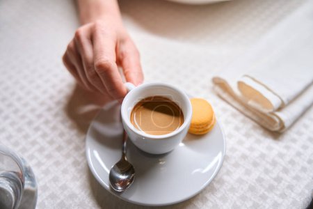 Foto de Taza de mano femenina de primer plano de café recién hecho fragante, disfrutando del sabor y el aroma de la bebida caliente y comiendo galletas sentadas en la cafetería - Imagen libre de derechos